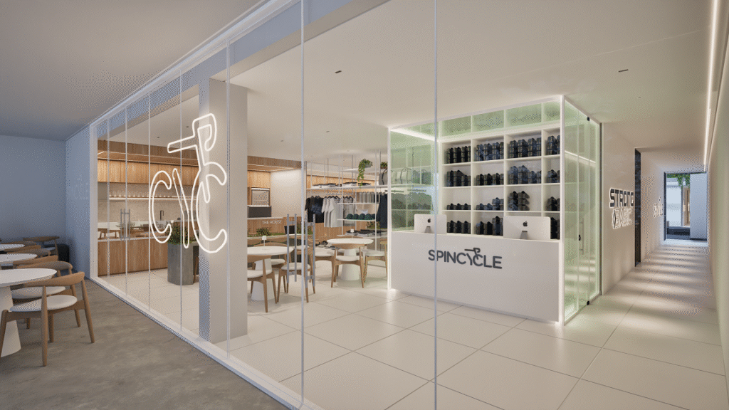 Sala moderna de academia boutique com decoração minimalista. Na frente, há um armário branco de prateleiras com muitos objetos organizados, provavelmente garrafas ou acessórios de ginástica, e a palavra "SPINCYCLE" estampada. Ao fundo, várias mesas e cadeiras de madeira clara compõem uma área de convivência. Há uma grande parede de vidro separando os ambientes, e, acima, uma luminária de neon com o desenho estilizado de uma bicicleta. O espaço é bem iluminado e transmite sensação de organização e modernidade.