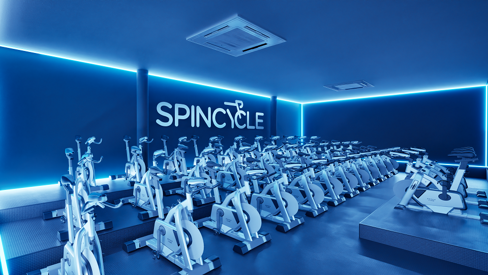 Sala de spinning com várias bicicletas ergométricas alinhadas em fileiras. As luzes do ambiente são em tons de azul neon, criando um clima moderno e energético. Na parede do fundo está escrito "SPINCYCLE" em letras grandes e brancas.