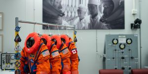 Four bright orange spacesuits with the circular NASA logo and American and Canadian flags on the arms hang on a rack in the middle of an empty room with chairs
