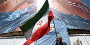 Homem-segura-bandeira-iraniana-perto-de-outdoor-anti-EUA-representando-o-presidente-dos-EUA-Donald-Trump-e-o-Estreito-de-Ormuz-em-Teera-Ira-em-30-de-maio-de-2026-e1782525884154