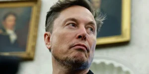 s Elon Musk is shown in the Oval Office, looking away from the camera to the right against a blurred backdrop showing paintings of former US presidents on the w