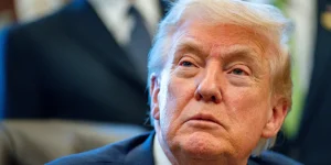 s Head and shoulders shot of Donald Trump in his office, looking upwards from his desk. He is wearing a blue jacket and white shirt