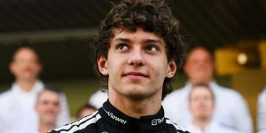 Kimi Antonelli como um piloto de corrida olhando para cima, com uma camisa de corrida preta, com pessoas atrás dele, embaçadas. Ele tem cabelo curto e encaracol