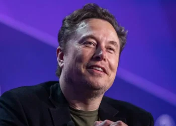 s Elon Musk stares towards the camera with a smile while sitting against a purple background on a stage