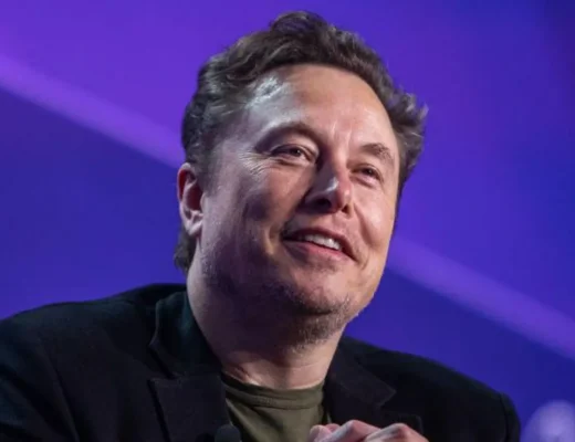 s Elon Musk stares towards the camera with a smile while sitting against a purple background on a stage