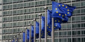 m colorida de bandeiras azuis com estrelas amarelas da União Europeia, dispostas uma ao lado da outra, tremulando, com um prédio ao fundo