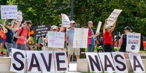 People hold signs outside on a sunny day in protest of NASA budget cuts, July 20, 2025